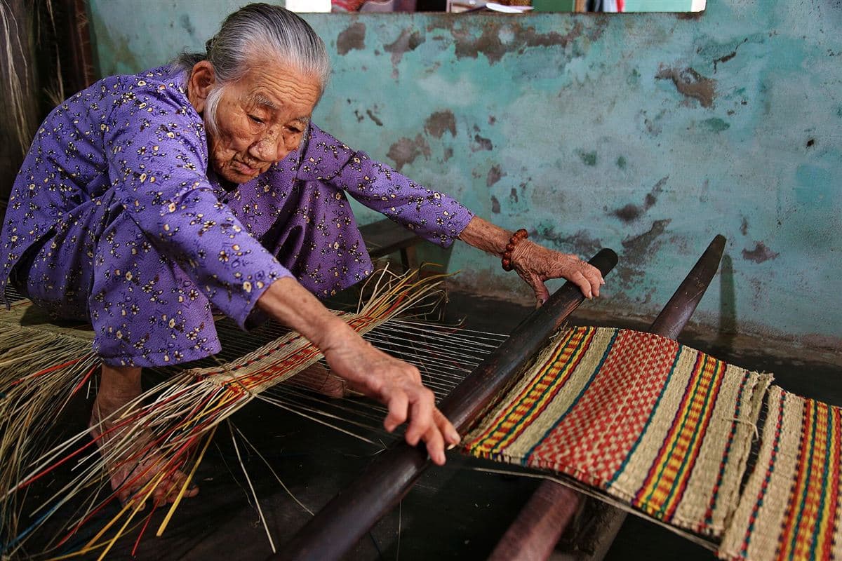 Cụ bà ngồi dệt chiếu cói hoa văn đỏ vàng bên khung dệt